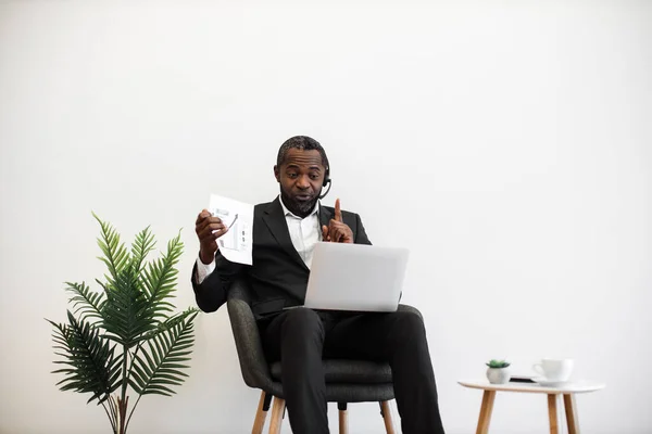 Competent african american financier presenting charts and graphs during online video meeting on modern laptop. Formally dressed man in headset sitting at office and having meeting on distance.