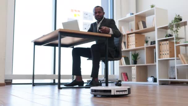 遥控机器人真空去除地板上的灰尘 而快乐的非洲商人在便携式计算机上工作 自动清洁改善工作间卫生 — 图库视频影像