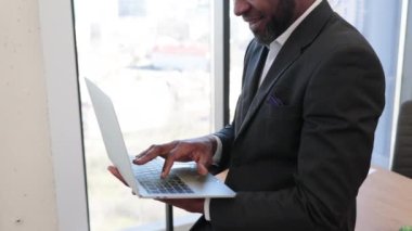 Elinde taşınabilir bilgisayarla ofis içi yazı masasının kenarında oturan çok kültürlü bir adama yakın durun. Verimli yaratıcı yönetici kablosuz bağlantı kullanarak e-ticaret web sayfalarına göz atıyor.