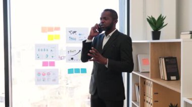 Takım elbiseli Afro-Amerikan bir adam rahat ofis odasında modern akıllı telefon kullanarak telefon görüşmesi yapıyor. Baş yönetici kablosuz teknoloji ile diğer liderlik yöneticileriyle iletişim kuruyor..