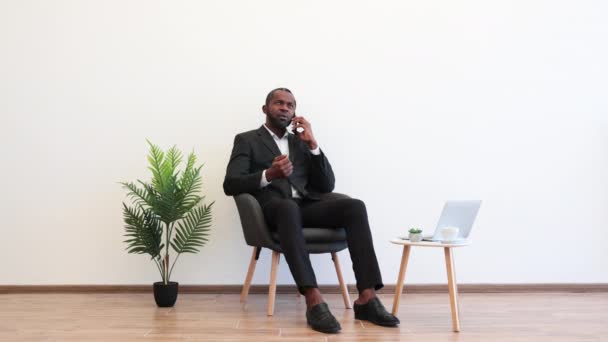 アフリカ系アメリカ人の男性は オフィスで現代的なスマートフォンを使いながら 顔の表情の広さを脇に置いています ラップトップとドリンクのカップとコーヒーテーブルの近くに座っているビジネススーツの男性雇用主 — ストック動画