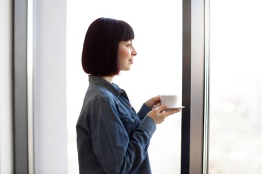 Beyaz kadın taze aromalı kahve içiyor ve panoramik pencereden manzaranın keyfini çıkarıyor. Bayan ofis çalışanının kahve molası..