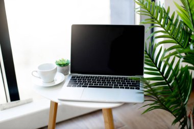 Kablosuz laptop, parlak ofisteki beyaz seramik bardağın yanındaki sehpanın üzerine yerleştirilmiş. Çalışanların rahat ve modern işyeri. İş için teknoloji kavramı.