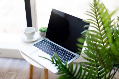 Kablosuz laptop, parlak ofisteki beyaz seramik bardağın yanındaki sehpanın üzerine yerleştirilmiş. Çalışanların rahat ve modern işyeri. İş için teknoloji kavramı.