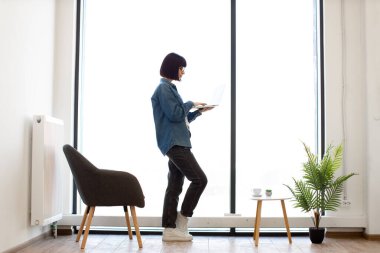 Ofisteki panoramik pencerenin yanında dururken kablosuz bilgisayarda yazı yazan çekici bir kadın. Koyu renk saçlı, günlük kıyafetli ve gözlüklü bir kadın iş yerindeki elektronik postaları kontrol ediyor..