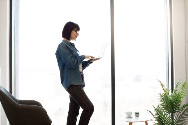Ofisteki panoramik pencerenin yanında dururken kablosuz bilgisayarda yazı yazan çekici bir kadın. Koyu renk saçlı, günlük kıyafetli ve gözlüklü bir kadın iş yerindeki elektronik postaları kontrol ediyor..