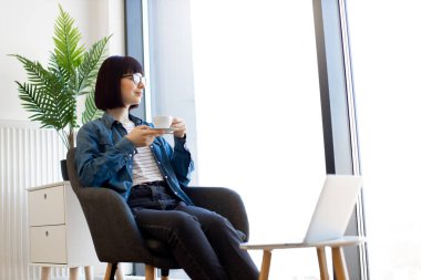 Elinde kahve fincanı olan sevimli bir kadın kablosuz bilgisayarı olan küçük bir masanın yanında oturuyor. Kafkasyalı esmer iş yerinde kahve molası sırasında panoramik pencereye bakarken sıcak içeceğin tadını çıkarıyor..