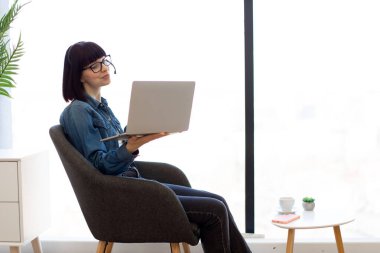 Elinde dizüstü bilgisayarla gözlüklü gülümseyen genç bir kadın akıllı telefonlu kahve masasının yanında oturuyor ve içiyor. Ofisteki kablosuz kulaklığı kullanarak çevrimiçi brifinge katılan etkili bir danışman..