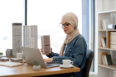 Başörtüsü takan neşeli müslüman kadın ve dizüstü bilgisayarda yazı yazan gözlüklü ofis masasında mimari bir model tutuyor. Becerikli şehir planlamacısı metropolitan alanında arazi kullanım programları geliştiriyor.