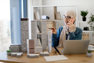 Mutlu Müslüman kadın tesettür ve gözlük takıyor. Ofis masasında modelleri tararken telefonla konuşuyor. Tecrübeli konsept mimarı takım üyelerine bilgi aktarıyor.