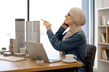 Ofisteki yenilikçi proje için taşınabilir bilgisayar kullanırken tesettürlü, tesettüre dönen neşeli Arap kadın. Yetenekli tasarımcı elinde kalemle ilham buluyor..