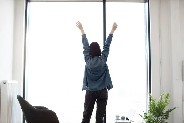 Stock image Back view of relaxed female brunette stretching arms while standing at spacious office near panoramic window. Caucasian young employee in casual attire enjoying coffee break during working hours.