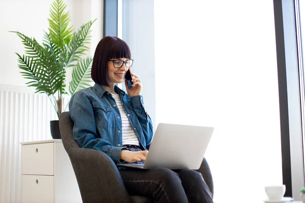 Happy caucasian woman in casual clothes talking on smartphone while surfing internet via laptop during coffee break in office. Business lady in eyeglasses using social networks during phone call.