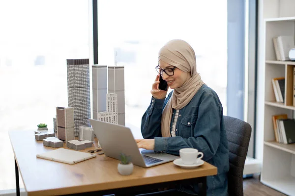 Усміхнена Мусульманка Хіджабі Повсякденному Одязі Розмовляє Смартфону Переглядаючи Інтернет Ноутбуком — стокове фото