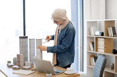 Ofiste mimari modellerle çalışırken bej tesettürlü, pusula kullanan mutlu Müslüman kadın. Verimli tasarım profesyonel ölçümleri bilgisayardaki dijital verilerle karşılaştırma.