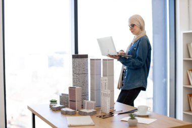 Mimari modellerle ofis masasının yanında dururken portatif bilgisayarda çalışan tesettür ve günlük giysiler içinde Müslüman bir kadın işçi. Yetenekli tasarımcı dizüstü bilgisayarda yeni proje planı oluşturuyor.