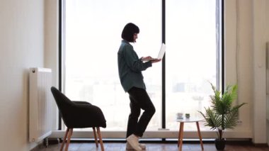 Ofisteki panoramik pencerenin yanında dururken kablosuz bilgisayarda yazı yazan çekici bir kadın. Koyu renk saçlı, günlük kıyafetli ve gözlüklü bir kadın iş yerindeki elektronik postaları kontrol ediyor..