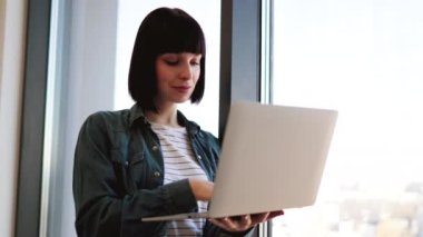 Parlak geniş bir ofiste çalışırken modern dizüstü bilgisayar kullanarak gündelik giysiler içinde odaklanmış esmer. Elinde taşınabilir cihazla panoramik pencerenin yanında duran beyaz kadın..