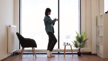 Kafkas esmer kadının tam boy portresi panoramik pencerenin yanında duruyor ve bileğindeki akıllı telefona bakıyor. Taşınabilir cihazda uygulamayı kullanarak günlük kıyafet giyen bir ofis çalışanı..