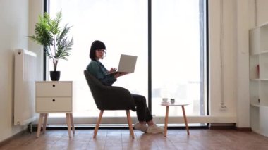 Kreatif iş yerinde sehpanın yanında otururken gözlüklü, modern dizüstü bilgisayarı tutan güzel bir kadın. Günlük kıyafetli neşeli iş kadını kablosuz kulaklıkla video konferansı düzenliyor..
