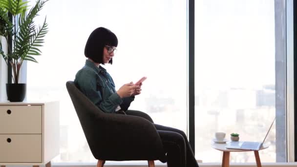 Mulher Cabelos Escuros Agradável Digitando Mensagens Smartphone Enquanto Sentado Cadeira — Vídeo de Stock