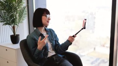 Kot ceketli genç beyaz kadın ofis içi kahve molasında selfie çubuğuyla cep telefonu kullanıyor. Çevrimiçi brifinge ev içi video başvurusuyla katılan etkin bir çalışan.