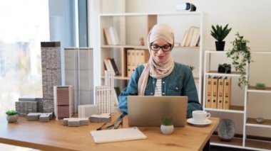 Gözlüklü, tesettürlü Müslüman kadın ofis masasındaki taşınabilir bilgisayarda modeller yazıyor. Deneyimli mimar modern aletler ve çizim araçları kullanarak şehir tasarımı geliştiriyor..