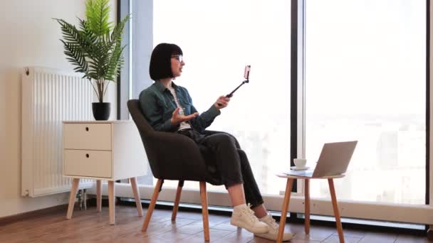 漂亮的年轻女人穿着牛仔夹克 一边拿着智能手机的自拍棒一边在办公室休息 一边喝咖啡 在舒适的工作环境中 微笑着带着胜利标志的女员工自画像 — 图库视频影像