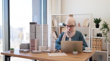 Yaratıcı iş yerinde kahve molası sırasında ofis masasında modellik yapan tesettürlü ve gözlüklü güzel bir Arap kadın. Mimari tasarımcı dizüstü bilgisayar kullanarak yeni projenin detaylarını analiz ediyor.