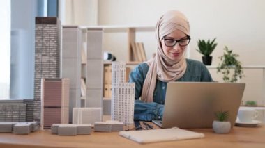 Yaratıcı iş yerinde kahve molası sırasında ofis masasında modellik yapan tesettürlü ve gözlüklü güzel bir Arap kadın. Mimari tasarımcı dizüstü bilgisayar kullanarak yeni projenin detaylarını analiz ediyor.