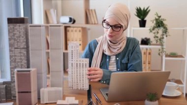 Modern çalışma alanında kentsel tasarım projesi üzerinde çalışırken dizüstü bilgisayarda bina modelini yöneten tesettürlü düşünceli Müslüman kadın. Deneyimli mühendis görüntülü arama uygulaması aracılığıyla çevrimiçi bilgilendirme yapıyor..