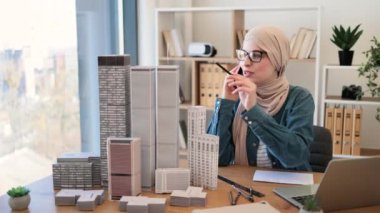 Tesettürlü, gözlüklü, tesettürlü, gülümseyen Müslüman bir kadın yaratıcı çalışma alanında ofis masasında otururken cep telefonuyla konuşuyor. Yetenekli mimar iş yerindeki müşterilerle iş görüşmesi yapıyor..