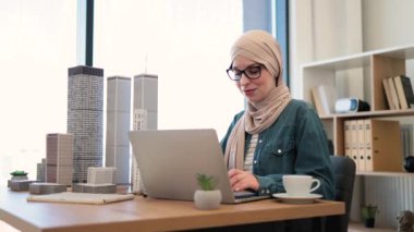 Gözlüklü, tesettürlü Müslüman kadın ofis masasındaki taşınabilir bilgisayarda modeller yazıyor. Deneyimli mimar modern aletler ve çizim araçları kullanarak şehir tasarımı geliştiriyor..