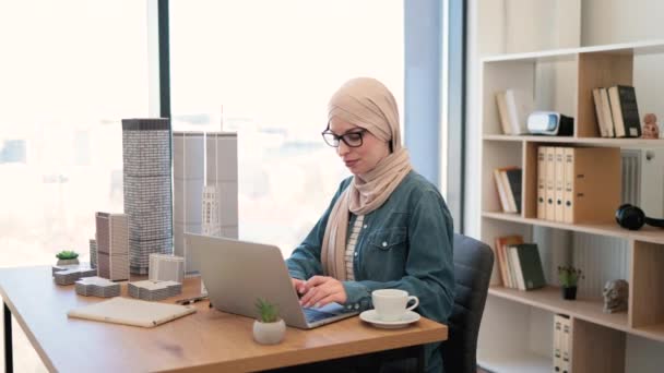 在写字台的手提电脑上用带眼镜的带头巾的穆斯林女性在打字 上面有建筑模型 有经验的建筑师利用现代小玩意和绘图工具开发城市设计 — 图库视频影像