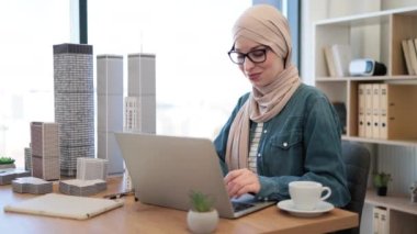 Başörtüsü takan neşeli müslüman kadın ve dizüstü bilgisayarda yazı yazan gözlüklü ofis masasında mimari bir model tutuyor. Becerikli şehir planlamacısı metropolitan alanında arazi kullanım programları geliştiriyor.
