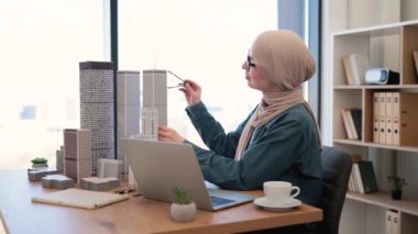 Tesettürlü, gözlüklü, tesettürlü, gülümseyen Müslüman bir kadın yaratıcı çalışma alanında ofis masasında otururken cep telefonuyla konuşuyor. Yetenekli mimar iş yerindeki müşterilerle iş görüşmesi yapıyor..
