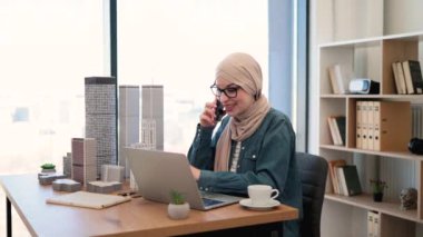Tesettürlü, gözlüklü, tesettürlü, gülümseyen Müslüman bir kadın yaratıcı çalışma alanında ofis masasında otururken cep telefonuyla konuşuyor. Yetenekli mimar iş yerindeki müşterilerle iş görüşmesi yapıyor..