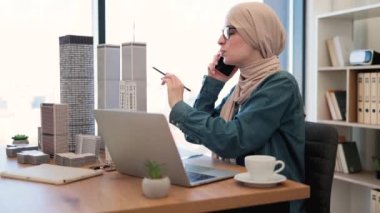 Kafiyeli kalem tutan ve mimarlık ofisinde modeller inşa ederken akıllı telefonla konuşan odaklanmış Arap kadın. Proje yöneticisi iş yerinde iş arkadaşlarıyla detayları tartışıyor