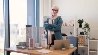 Yetişkin Arap kadın tesettürlü yazı masasının arkasında, mimarlık ofisinde aletler ve maketlerle poz veriyor. Yaratıcı iş yerindeki proje yönetimine kahve molası veren deneyimli bir profesyonel..