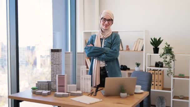 建築事務所でガジェットやマケットを机の後ろにポーズをヒジャーブ語で大人のアラビア語の女性 創造的な職場でのプロジェクト管理からコーヒーブレイクを持つ経験豊富な専門家 — ストック動画