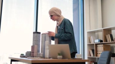Şehir kompleksinin mimari modellerini dizayn eden tesettürlü ve gözlüklü olumlu Arap bayan dizüstü bilgisayar ve tabletin yanındaki ofis masasına yerleştirildi. Yetenekli profesyonel kentsel alan projesi iş başında.