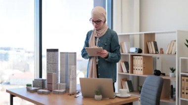 Bej tesettürlü gülümseyen Arap kadın ofis masasındaki mimari modellerin yanına dizüstü bilgisayarla dijital tablet üzerinde çalışıyor. Teknolojileri kullanarak profesyonel inşaat projesi tasarla.