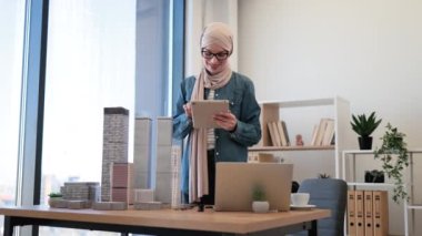 Türbanlı neşeli müslüman kadın ve modern tablet taşıyan gözlüklü kadın yazı masasından çok katlı gökdelenin ölçekli modelini alırken. Mutlu tasarımcı yaratıcı iş yerinde dijital taslak denetimi yapıyor.