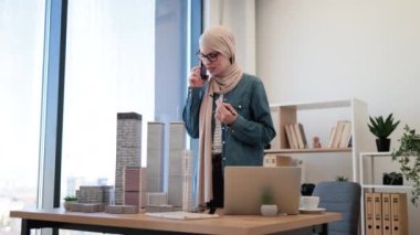 Bej tesettürlü gülümseyen Arap kadın bina modelinde kalemle el ele tutuşurken yaratıcı iş yerinde telefonla konuşuyor. Mimarlık ofisinde iş konuşması yapan yetenekli bir profesyonel..