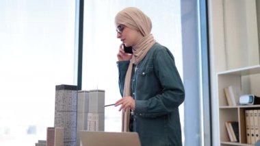 Bej tesettürlü Arap iş kadını ofiste yazı masasındaki modelleri tararken cep telefonuyla sohbet ediyor. Şehir kompleksi projesiyle ilgili iş arkadaşıyla beyin fırtınası yapan deneyimli tasarımcı.