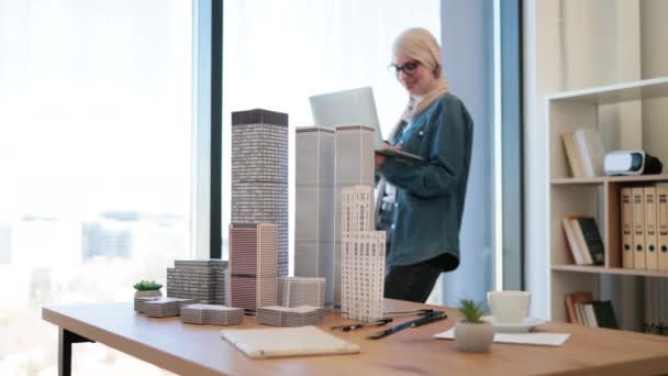 創造的な職場でパノラマの窓の近くに手にラップトップで立ってヒジャーブとメガネを身に着けているアラビアの実業家 ガジェットを使用して都市複合建設計画を実行する若い建築家 — ストック動画