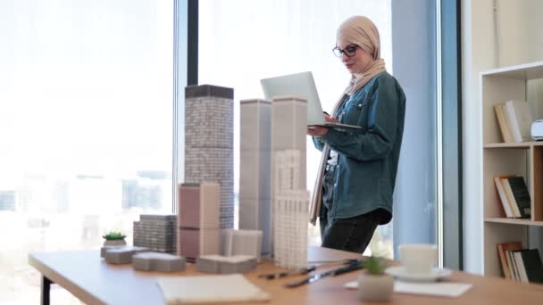 在创意工作场所 阿拉伯女商人手握手提电脑 头戴头巾 手握眼镜 站在全景窗前 利用小工具实施城市综合建设计划的年轻建筑师 — 图库视频影像