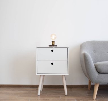 Endüstriyel edison lambası, geniş stüdyo odasındaki gri kanepenin yanındaki beyaz komodinin üzerinde. Beyaz ve gri renklerin minimalist iç kısımlarında loş, sıcak bir ışık sağlar..
