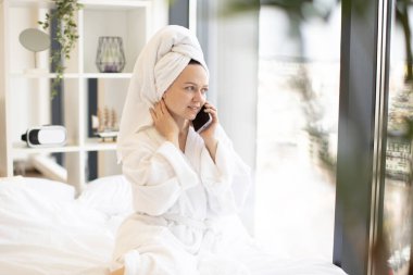 Beyaz bornozlu çekici beyaz kadın geniş yatak odasının panoramik penceresinden dışarı bakarken akıllı telefonla konuşuyor. Serene Lady kablosuz bağlantı kullanarak romantik bir akşam partisi planlıyor..