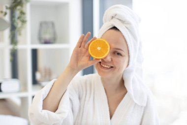 Beyaz bornozlu çekici sakin bir kadın ve başının üzerinde havlu. Bir gözü turuncu, diğer yarısı da odanın arka planında bulanık. Rahatlamış kadın güzellik için C vitamini açısından zengin narenciye uyguluyor..